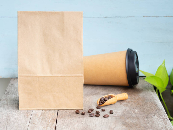 Pražená kávová zrna v papírovém sáčku s dřevěnou kopečkou, šálek na dřevěném stole. Mouckup, šablona s kopírovacím prostorem pro text nebo logo - Fotografie, Obrázek
