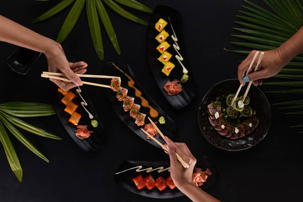 Top view hands with chopsticks and mix of sushi, Japanese food over black table with tropical leaves - Photo, image