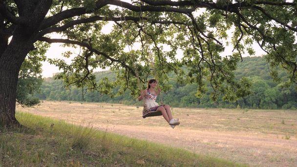 Una chica adulta balanceándose en un columpio atado a un árbol en la naturaleza, rodando en el aire en el viento y riendo, el concepto de una vida feliz - Foto, Imagen