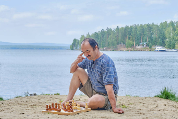 Homem sênior ponderando um movimento em um jogo de xadrez e sentado na praia perto do lago - Foto, Imagem