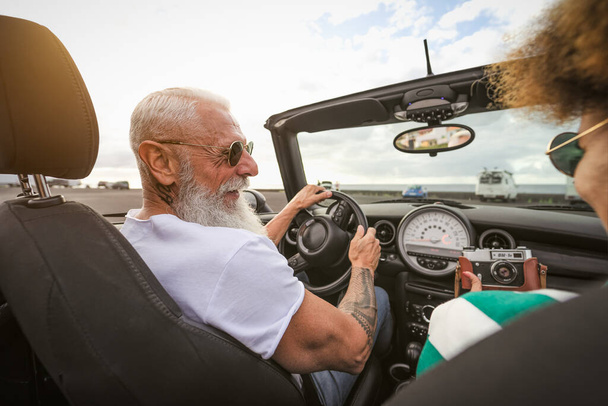 Feliz pareja de ancianos que se divierten tomando fotos con la vieja cámara vintage mientras conducen en un coche nuevo convertible durante las vacaciones de viaje por carretera - Foto, Imagen