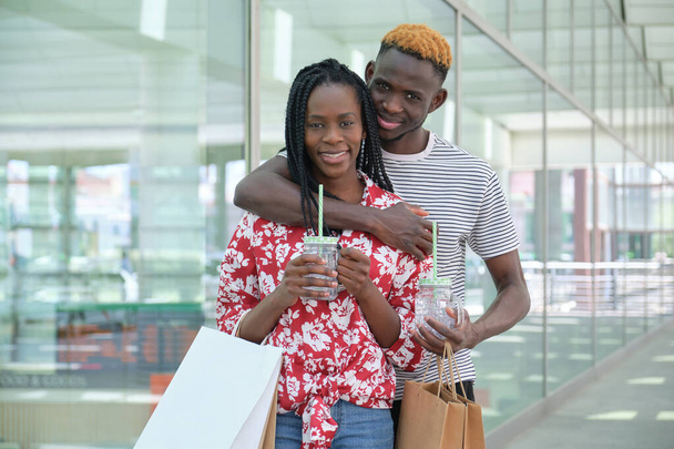 Portret młodej afrykańskiej pary pijącej orzeźwiające drinki i idącej na zakupy, przytulającej się i patrzącej w kamerę. Koncepcja życia miasta. - Zdjęcie, obraz