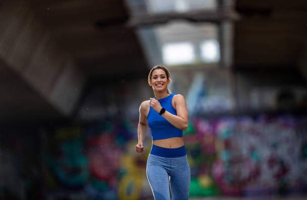 Sportler läuft lächelnd vor Mauer mit Graffiti. Mädchen mit Sportkleidung beim Sport. - Foto, Bild