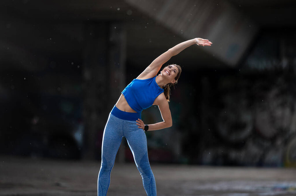 Atleta feminina se aquecendo esticando na frente da parede de grafite. Menina vestindo sportswear exercício sorrindo.  - Foto, Imagem