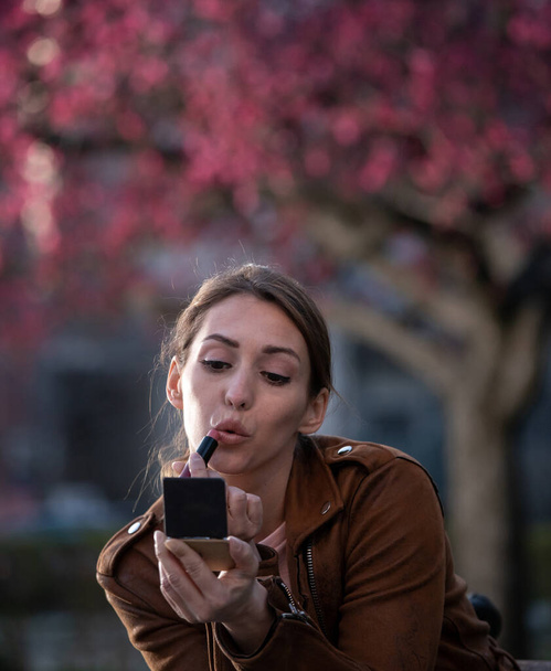 Studentin trägt Lippenstift im Park auf. Junge attraktive Frau benutzt im Frühling Schminkspiegel draußen.  - Foto, Bild