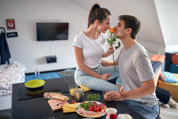 A young couple in love is smelling a rose while having a breakfast on Valentine's day in a romantic atmosphere at home together. Love, together, breakfast, home - Foto, imagen