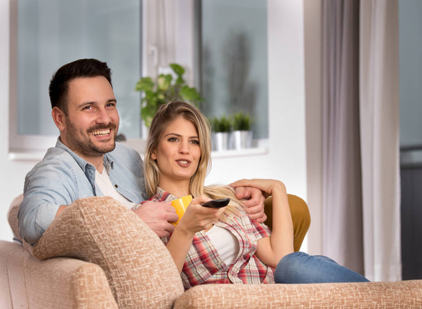 Jovem mulher segurando remoto assistindo tv com homem feliz sorridente. Casal sentado juntos na sala de estar.  - Foto, Imagem
