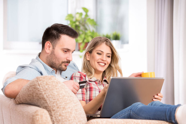 Junges Paar beim gemeinsamen Einkaufen im Wohnzimmer mit Laptop und Kreditkarte. Mann und Frau lächeln glücklich und zeigen auf Bildschirm. - Foto, Bild
