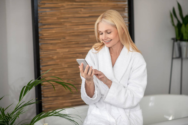A woman in a white bath robe with a smartphone in her hands - Valokuva, kuva