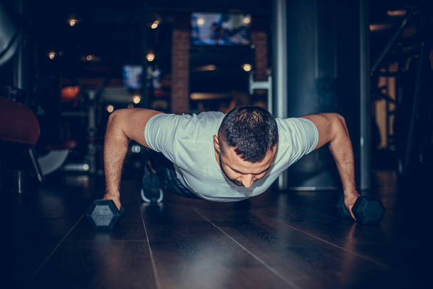 Man doing workout with heavy dumbbell. Confident muscled young man wearing sport wear and doing plank position while exercising on the floor in loft interior. - Foto, imagen