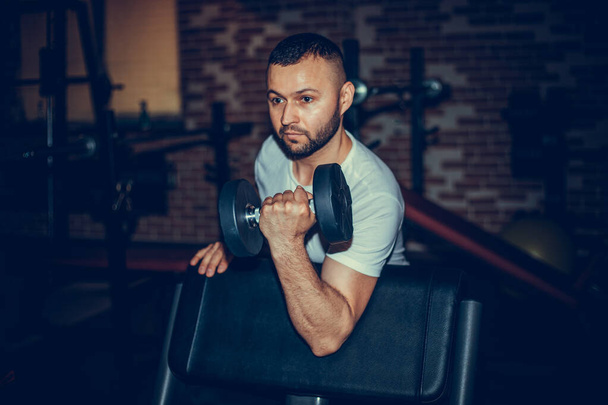 Fit a svalnatý muž dělá biceps cvičení s činkami v tmavé tělocvičně. - Fotografie, Obrázek
