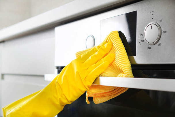 Cleaning service or housework. Female hand in a yellow rubber glove wipes built-in oven in a kitchen facade with microfiber. - Фото, изображение
