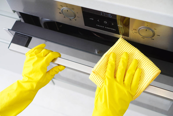 Cleaning service or housework. Female hand in a yellow rubber glove wipes built-in oven in a kitchen facade with microfiber. - Фото, зображення