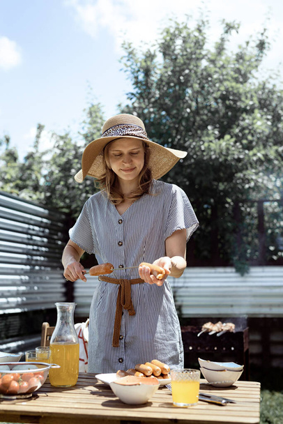 Young woman in summer hat and dress grilling meat and vegetables outdoors in the backyard. - Fotoğraf, Görsel