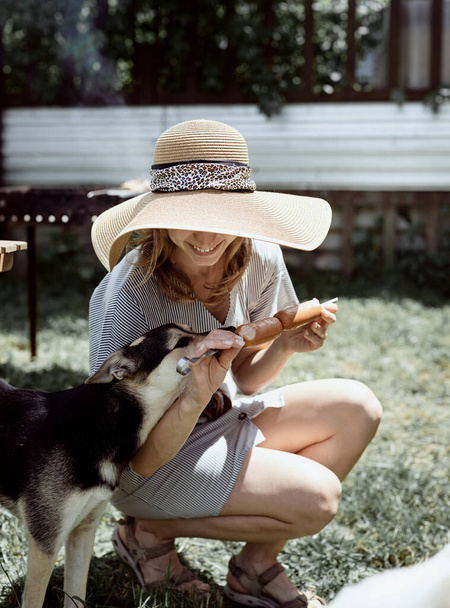 Jonge vrouw in de zomer hoed grillen vlees buiten in de achtertuin, zitten met haar hond, geven huisdier een snack - Foto, afbeelding