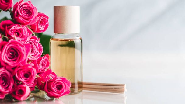 Clear bottle of yellow oil or perfume with wooden sticks and delicate pink roses on white surface. SPA salon and procedures concept. Banner. Copy space - Photo, Image