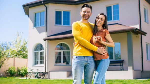 Modern beautiful excited young couple in love standing in front of the new big house with the keys in hands and hugging while they celebrating this buying - Photo, image