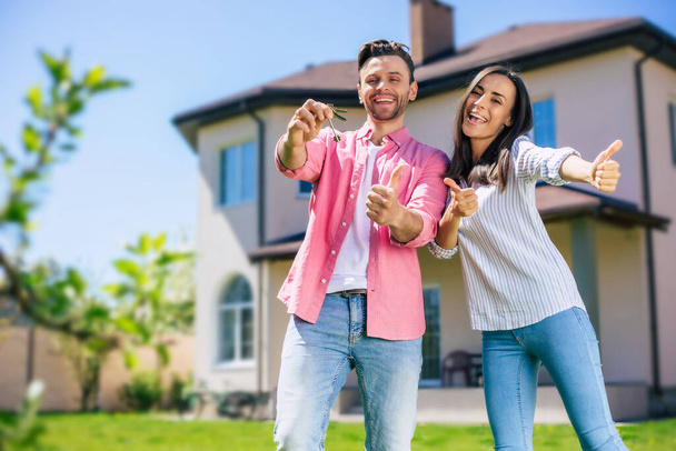 Modern beautiful excited young couple in love standing in front of the new big house with the keys in hands and hugging while they celebrating this buying - Foto, Bild
