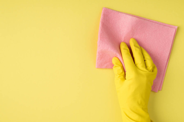 Top view photo of hand in yellow rubber glove using viscose napkin on isolated yellow background with copyspace - Foto, afbeelding
