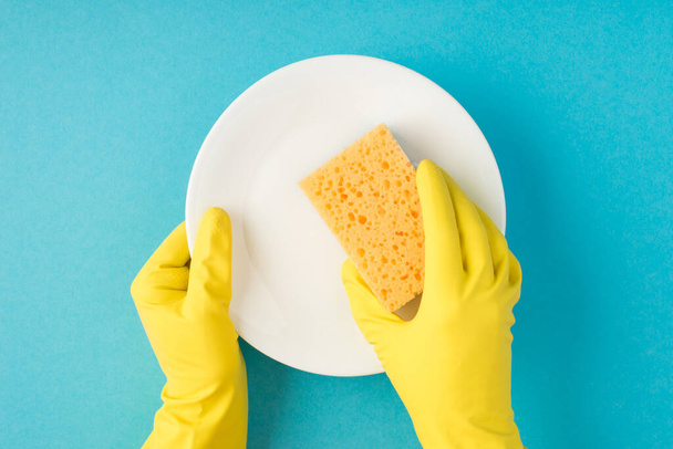 Vista superior foto de las manos en guantes de goma amarilla sosteniendo esponja amarilla y plato blanco limpio sobre fondo azul pastel aislado - Foto, imagen