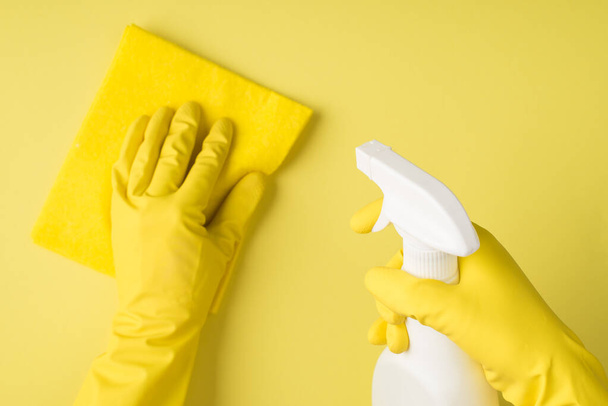 Bovenaanzicht foto van handen in gele rubberen handschoenen met spray wasmiddel witte fles en viscose doek op geïsoleerde gele achtergrond met copyspace - Foto, afbeelding
