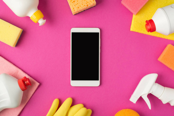 Фото смартфона в центре прочесывания лохмотья резиновые перчатки и белые бутылки моющего средства на изолированном розовом фоне с копипространством - Фото, изображение