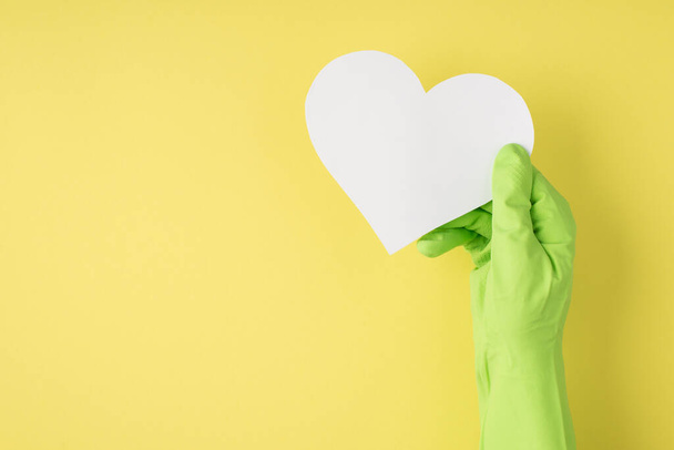Foto vista superior de la mano en guante de goma verde sosteniendo el corazón de papel blanco sobre fondo amarillo aislado con espacio en blanco - Foto, imagen