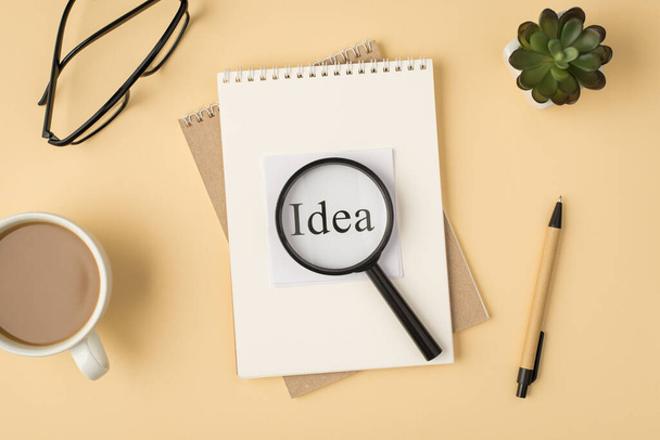 Top view photo of sticker with world idea magnifier on two notebooks pen glasses plant and cup on isolated beige background - 写真・画像
