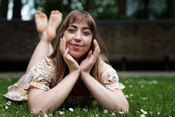 Retrato de una joven tumbada boca abajo sobre la hierba en un día de primavera. Lleva un vestido de primavera y sonríe.. - Foto, Imagen