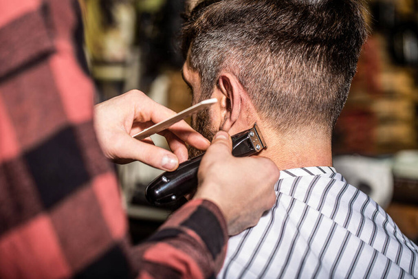 Mężczyzna odwiedzający fryzjera w salonie fryzjerskim. Fryzjer pracuje z maszyną do strzyżenia. Hipsterski klient strzyże się. Ręce fryzjera z maszynką do strzyżenia, zbliżenie. Koncepcja fryzury. Hipsterski klient obcina włosy - Zdjęcie, obraz