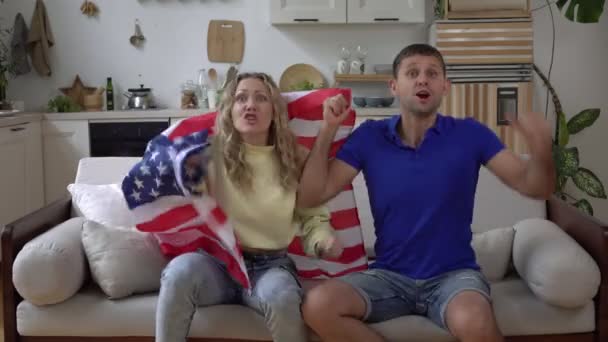 Семейная пара футбольных фанатов дома с флагом США. Американские болельщики смотрят телевизор дома на диване - Кадры, видео