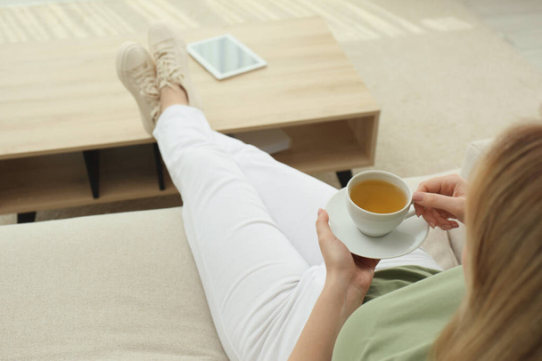 Young woman with cup of hot tea relaxing on sofa at home, closeup - Foto, afbeelding