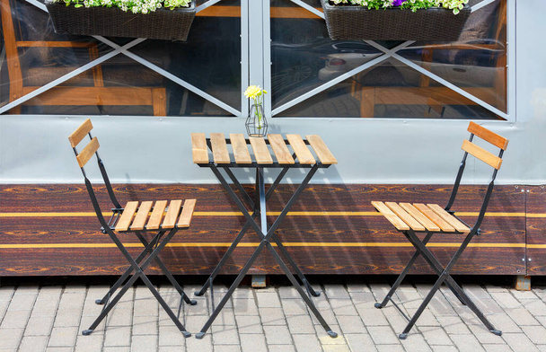 Yazlık kafenin kaldırımında bahçe mobilyaları, katlanabilir masa ve şehrin arka planında sandalyeler ve sepetlerde çiçekler var.. - Fotoğraf, Görsel
