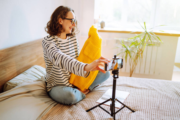 Fashion blogger. Young woman blogger talking with followers, live streaming and showing yellow sweater on camera. Stylist influencer woman  showing trendy clothes filming vlog episode for her channel. - Foto, afbeelding