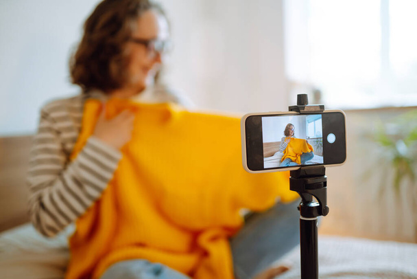 Fashion blogger. Young woman blogger talking with followers, live streaming and showing yellow sweater on camera. Stylist influencer woman  showing trendy clothes filming vlog episode for her channel. - Photo, image