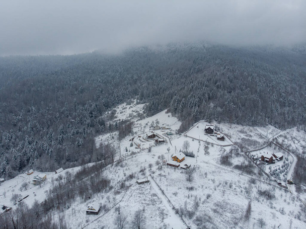 Luftaufnahme eines Bergdorfes mit schneebedeckten Hügeln und Kiefernwald im Winter. Jaremtsche, Ukraine. Fliegen über die verschneiten Hügel und hölzernen Häuschen. Land, Tanne. Drohnen-Ansicht - Foto, Bild