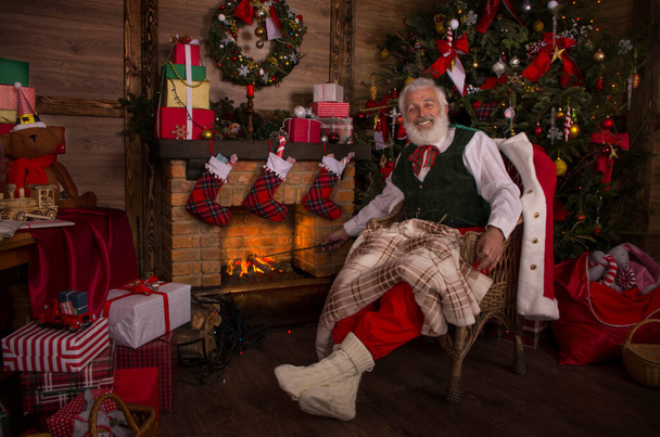Санта Клаус біля каміна і ялинки. Святковий інтер'єр всередині дерев'яного будинку, веселий настрій Нового року Дух Різдва. Старша людина з справжньою білою бородою косплей Батько Різдво. - Фото, зображення