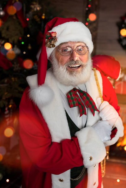 暖炉とクリスマスツリーの近くのプレゼント付きのバッグ付きサンタクロース。木製の家の中のお祭りのインテリア、クリスマスの新年の陽気な気分スピリット。シニア男とともに本当の白いひげのコスプレ父クリスマス. - 写真・画像