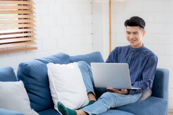 Jeune homme d'affaires asiatique sourire et travailler de la maison avec ordinateur portable en ligne à Internet sur le canapé dans le salon, homme indépendant en utilisant un ordinateur portable sur le canapé avec confort, nouveau concept normal, style de vie. - Photo, image