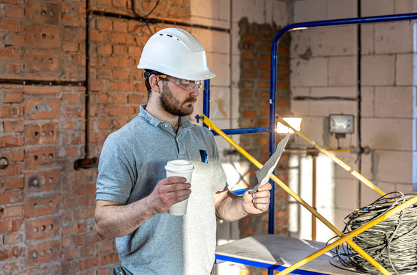 Bir elektrikçi elinde kahveyle bir inşaat çizimini inceliyor.. - Fotoğraf, Görsel
