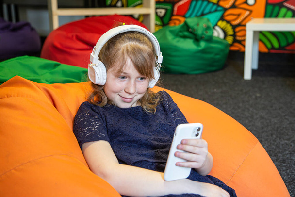 Little girl uses phone and listens to music with headphones while sitting on bag chair. - Photo, Image