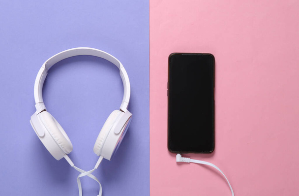 紫ピンクのパステルカラーの背景にスマートフォンに接続されたステレオヘッドフォン - 写真・画像