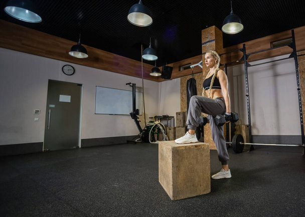 Η Fit woman στο χώρο του αθλητισμού εξασκείται σε lunges με αλτήρες στα χέρια της σε ένα ξύλινο κουτί σε ένα σύγχρονο γυμναστήριο. Λειτουργική εκπαίδευση - Φωτογραφία, εικόνα