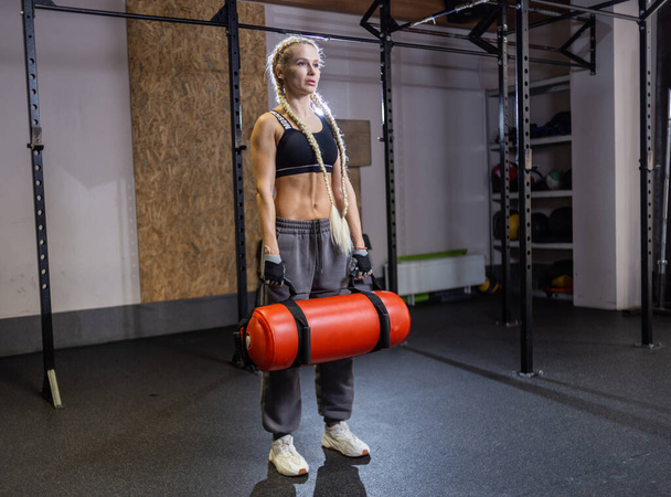 Спортивная сильная женщина упражняется с тяжелой функциональной тренировочной сумкой в современном тренажерном зале  - Фото, изображение
