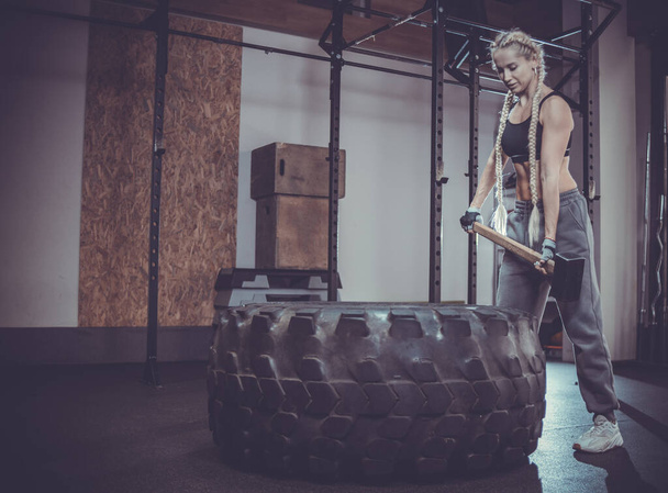 Muscular woman in sports underwear holding sledgehammer near tire wheel in modern gym - Photo, Image
