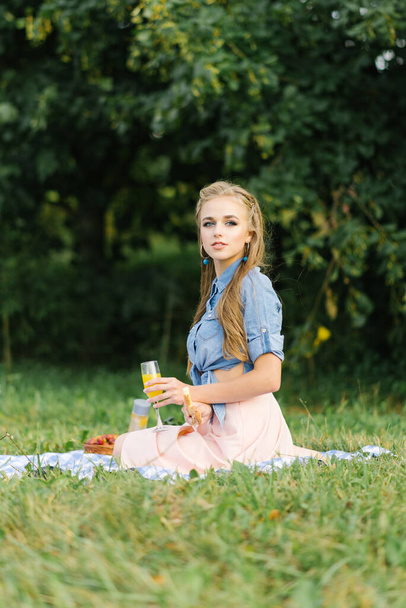 Uśmiechnięta piękna dziewczyna trzyma świeżą bułkę w rękach i siedzi na kocu na świeżym powietrzu w parku. Letni piknik w parku - Zdjęcie, obraz