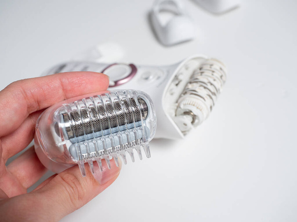Close-up van een hand die een mondstuk voor een witte elektrische epilator op een witte achtergrond vasthoudt. Op de achtergrond de epilator zelf en andere bijlagen. Schoonheidsmiddel - Foto, afbeelding