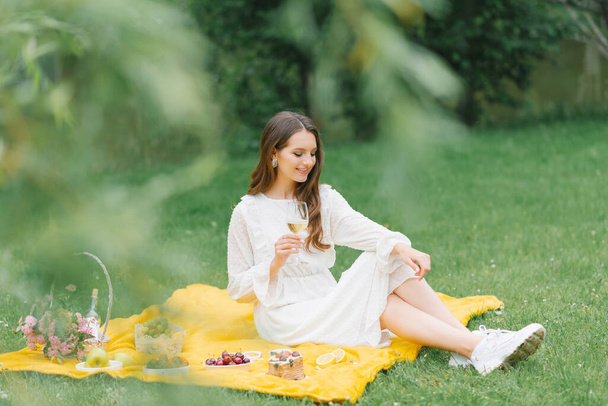 Mulher bonita em um vestido branco bebendo vinho no jardim em um piquenique - Foto, Imagem