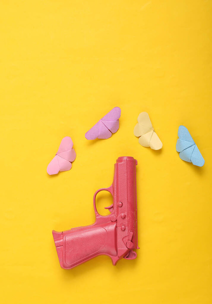 Розовый пистолет с бабочками на желтом фоне. Творческая концепция. Минимализм лежал. Мбаппе. - Фото, изображение