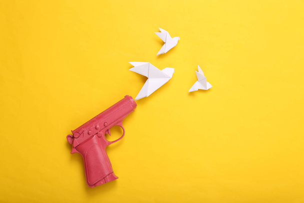 Pinkfarbene Pistole, die Origami-Tauben auf gelbem Hintergrund schießt. Kein Krieg. Kreative minimalistische Gestaltung. Flach lag er. Ansicht von oben - Foto, Bild